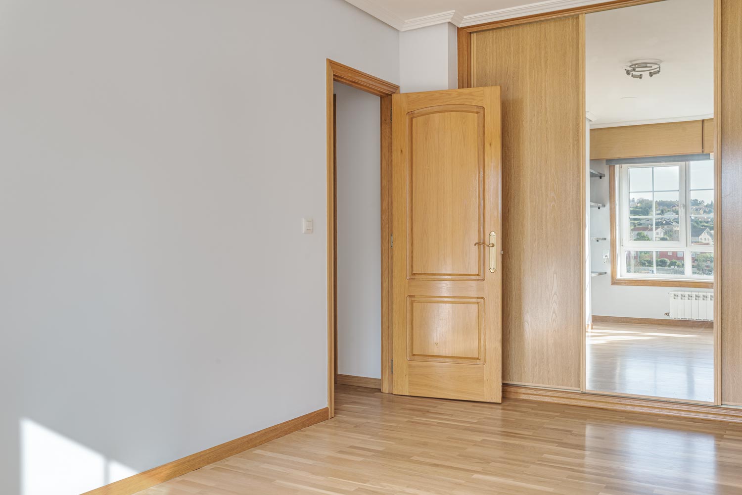 Vista de una habitación vacía con suelo de parqué claro y una puerta abierta de madera que muestra otra habitación iluminada con luz natural