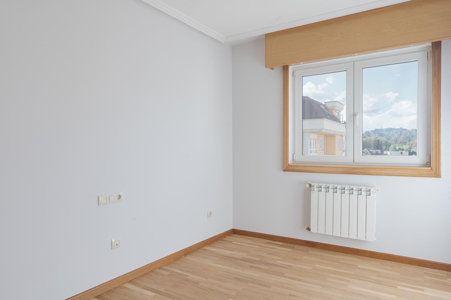 Una habitación vacía con suelo de parqué claro, una ventana de madera que muestra vistas exteriores y un radiador blanco, con paredes en tono gris claro