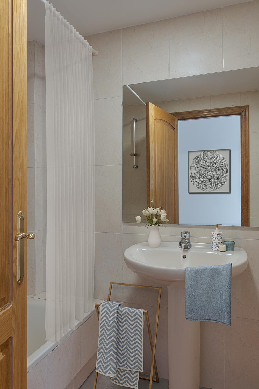 Baño con un lavabo de pedestal, un espejo con marco de madera, una bañera con cortina blanca y detalles decorativos como flores y una toalla de mano con patrón de chevron en un toallero de madera