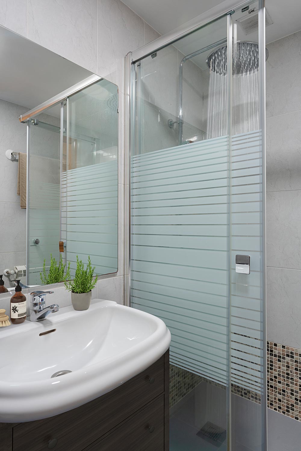 Baño contemporáneo con lavabo sobre mueble de madera oscura, accesorios de baño, una planta pequeña y una cabina de ducha con puertas de vidrio y azulejos claros.