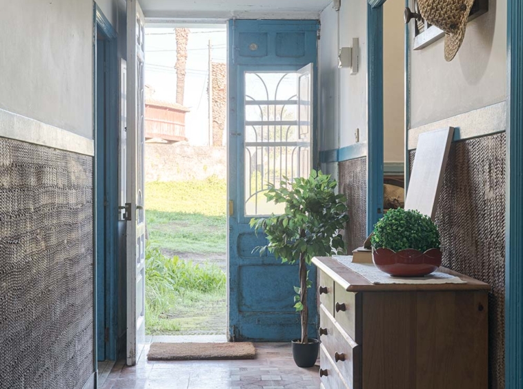 Entrada de casa con puerta azul abierta hacia un jardín verde, mostrando un pasillo acogedor con iluminación natural y detalles rústicos