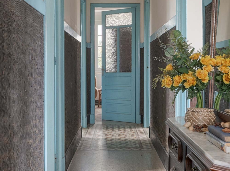 Pasillo largo con puertas azules, suelo de baldosas y espejos que reflejan un arreglo floral, aportando un aire rústico y acogedor en una casa de Bergondo, A Coruña