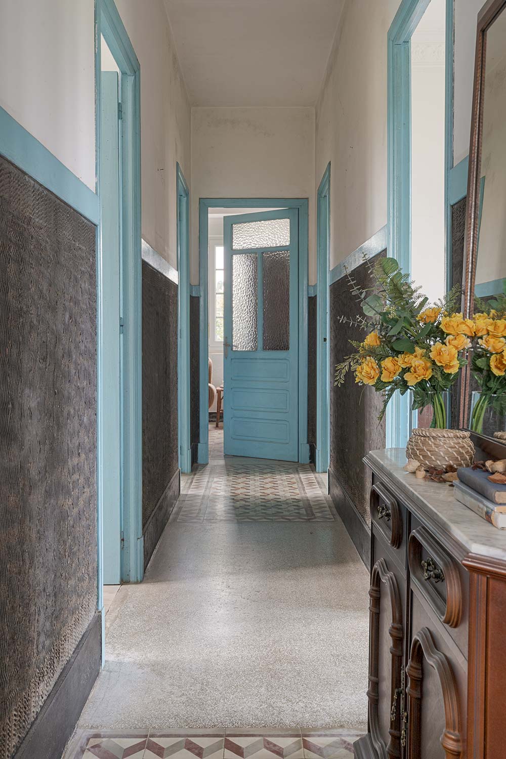 Pasillo largo con puertas azules, suelo de baldosas y espejos que reflejan un arreglo floral, aportando un aire rústico y acogedor en una casa de Bergondo, A Coruña