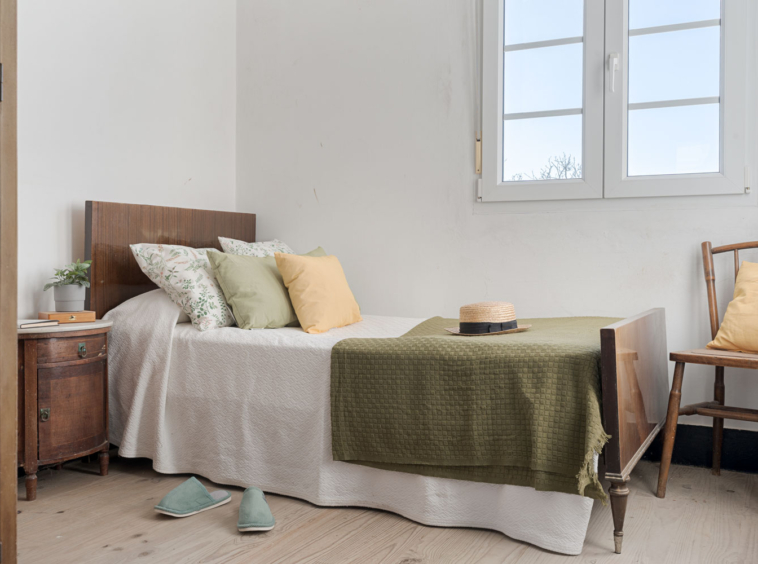 Habitación sencilla y bien iluminada con cama de madera, ropa de cama en tonos suaves y un sombrero de paja sobre la colcha, en una casa de Bergondo, A Coruña