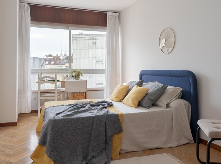 Dormitorio principal en un piso en Sada, A Coruña, decorado con técnicas de home staging, destacando una cama con cabecero azul y cojines amarillos y grises