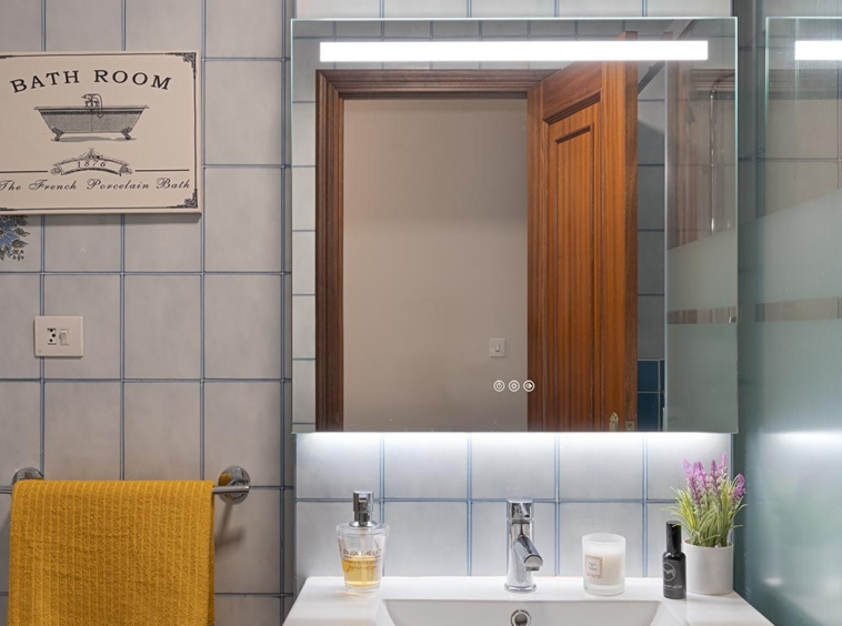 Baño con azulejos grises, un amplio espejo iluminado y detalles en madera, complementado con una toalla amarilla decorativa en un piso en Sada, A Coruña.