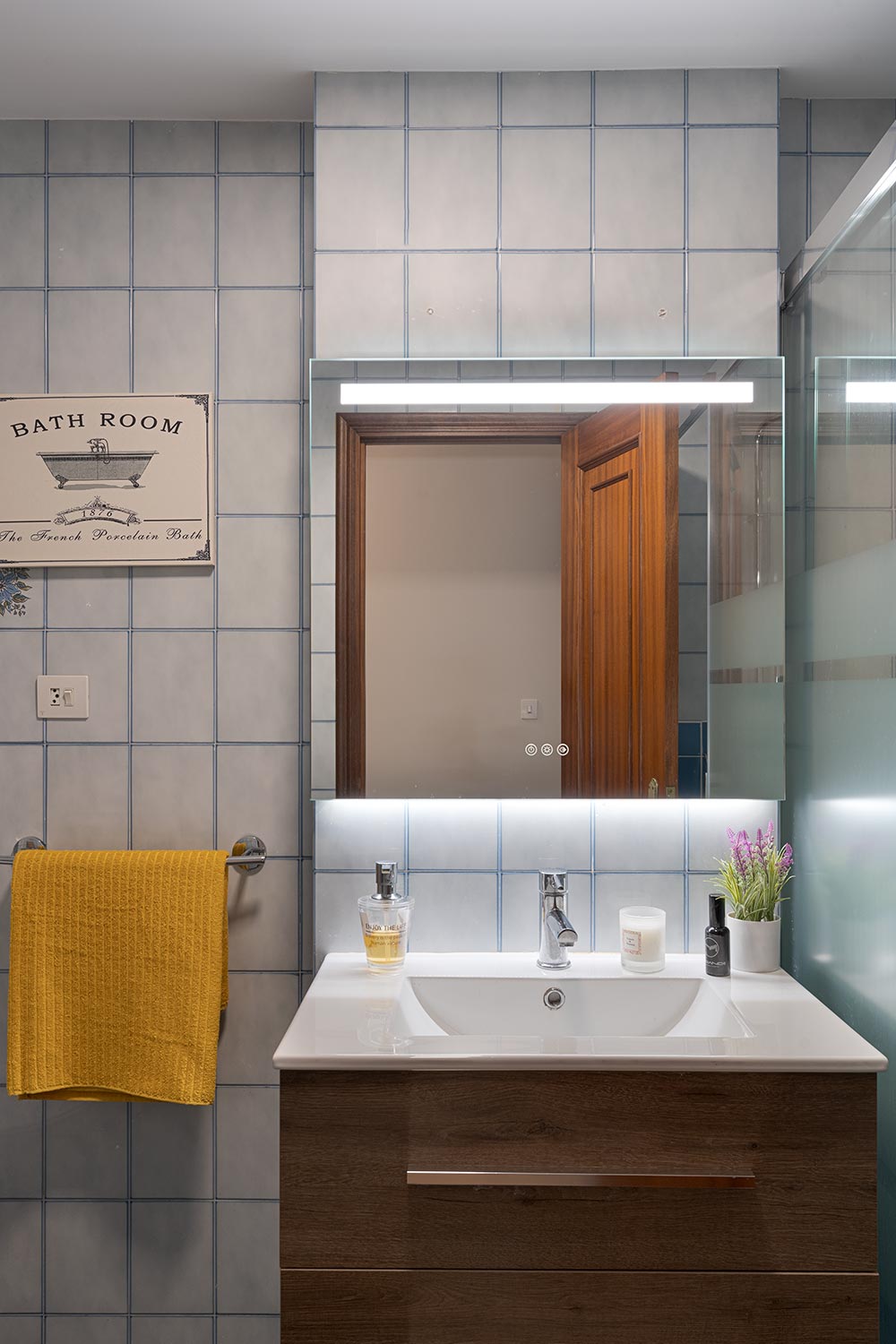 Baño con azulejos grises, un amplio espejo iluminado y detalles en madera, complementado con una toalla amarilla decorativa en un piso en Sada, A Coruña.