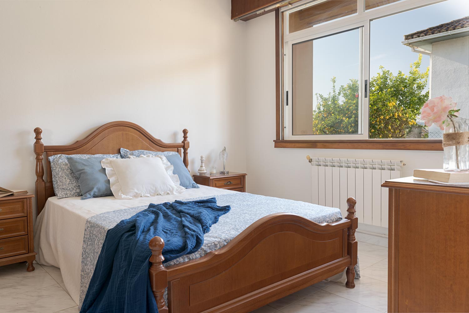 Dormitorio luminoso con cama de madera, ropa de cama en tonos azules y ventana con vistas al jardín.