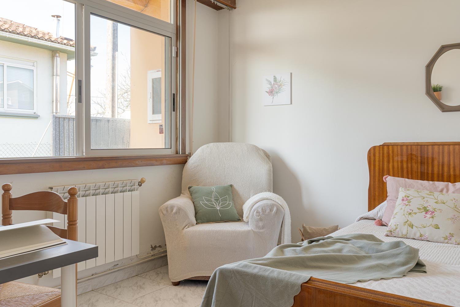 Habitación con cama sencilla, sillón cubierto con funda blanca y ventana que deja entrar luz natural.