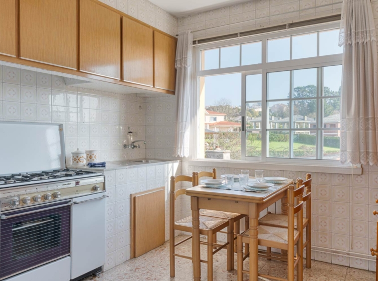 Cocina con mobiliario de madera clara, encimera de azulejos blancos y ventana grande con vistas al jardín.