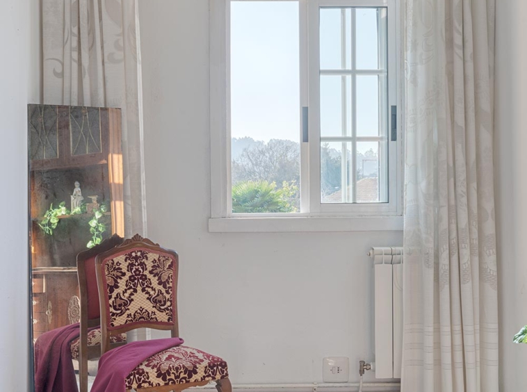 Ventana luminosa con cortinas y silla clásica con tapizado rojo y blanco en una habitación.