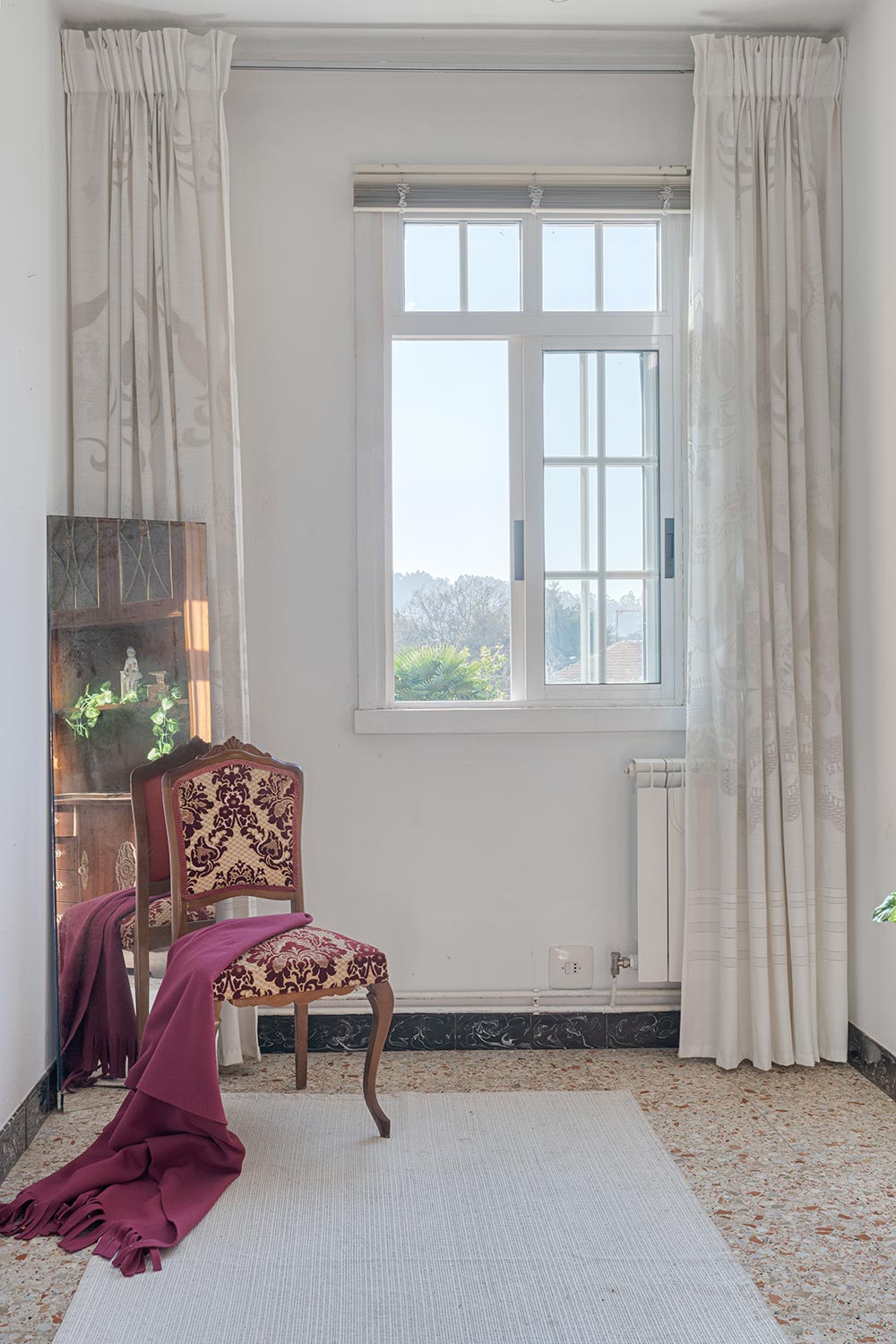 Ventana luminosa con cortinas y silla clásica con tapizado rojo y blanco en una habitación.