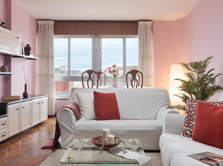 Sala de estar con paredes rosas, sofás blancos con cojines rojos, y mesa de comedor con vista a la ventana