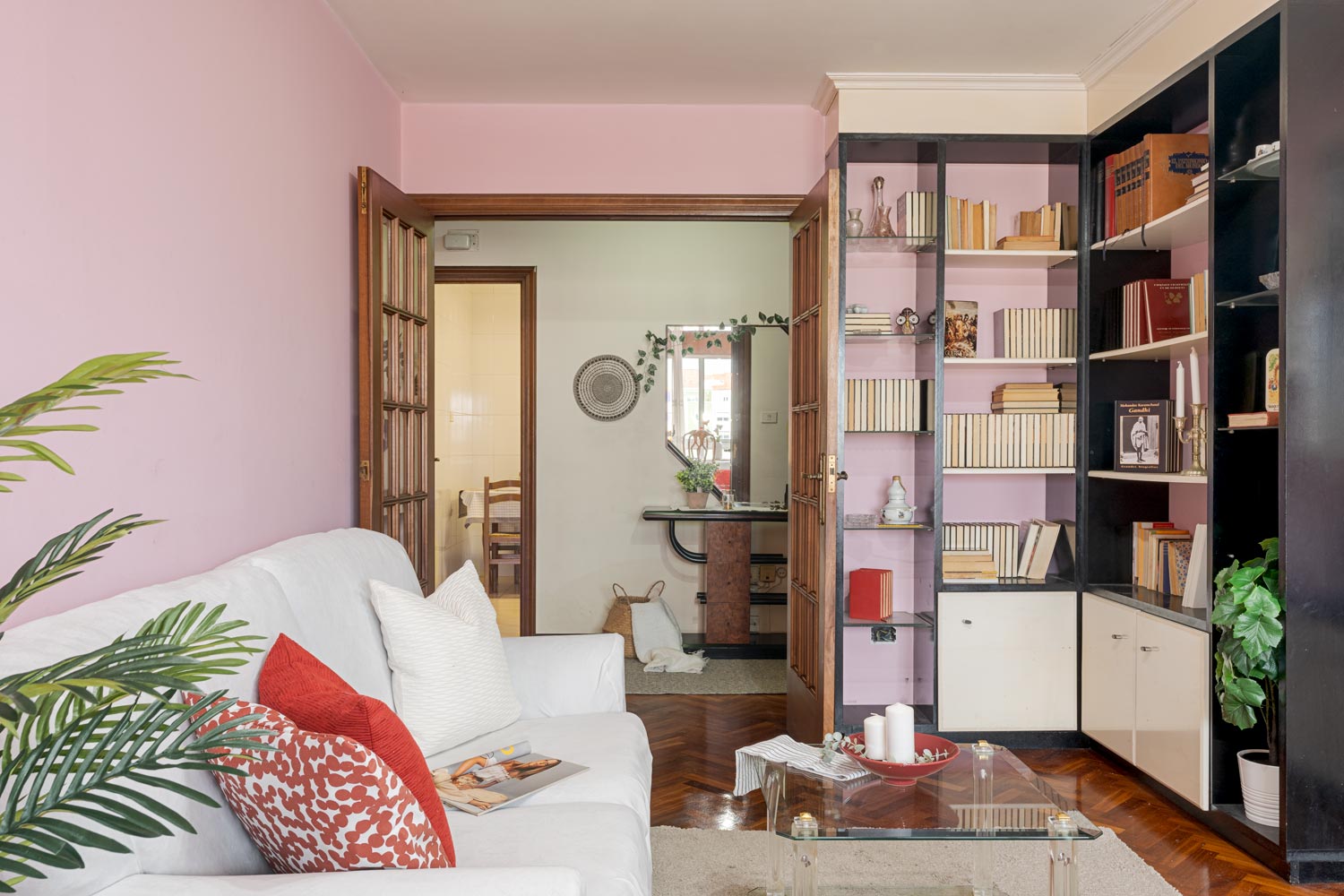 Salón con sofás blancos, cojines rojos, una estantería llena de libros y puertas de madera que conducen a la entrada