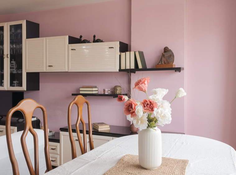 Comedor con mesa cubierta por mantel blanco, sillas de madera curvada, y una decoración de florero blanco con flores de colores sobre fondo de pared rosa