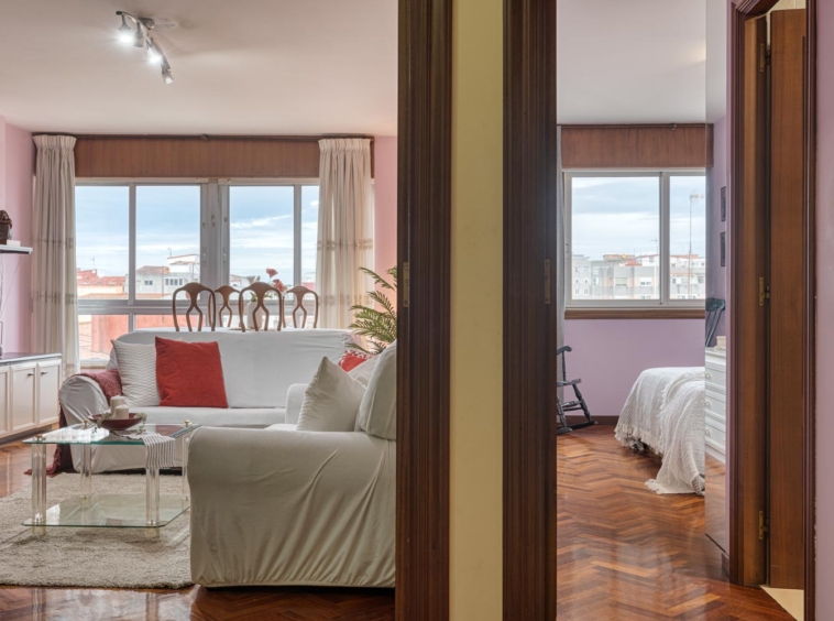 Espacioso salón y dormitorio con grandes ventanas que ofrecen luz natural, combinando tonos de rosa en las paredes y cojines con un sofá blanco y mesa de centro de vidrio