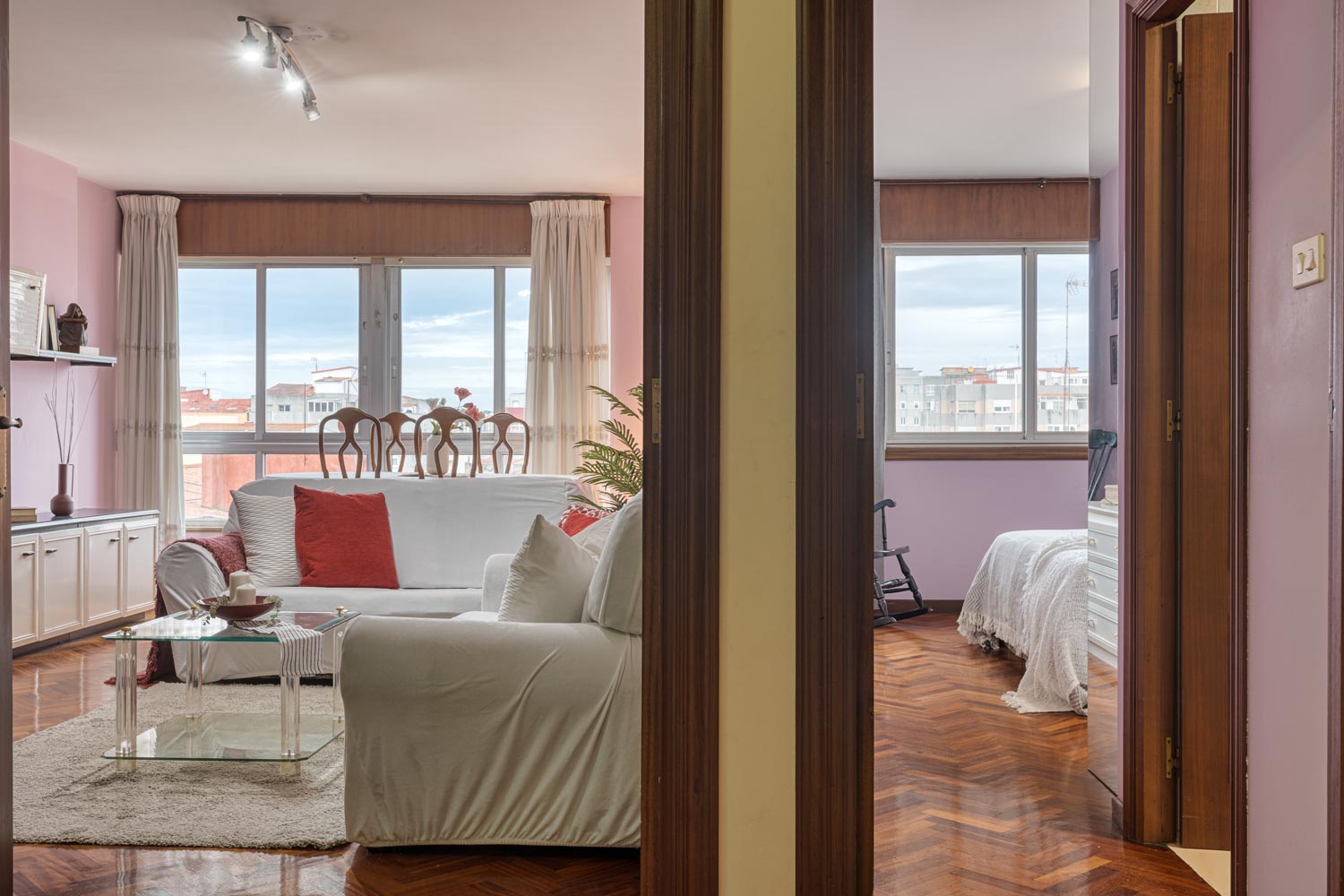 Espacioso salón y dormitorio con grandes ventanas que ofrecen luz natural, combinando tonos de rosa en las paredes y cojines con un sofá blanco y mesa de centro de vidrio