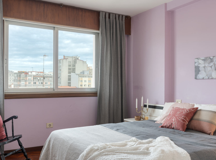 Dormitorio íntimo con paredes de un suave tono lavanda, gran ventana que permite la entrada de luz y ofrece vistas urbanas, decorado con una cama cubierta con textiles en tonos grises y crema