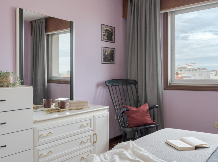 Acogedor rincón de un dormitorio con decoración en tonos lavanda y gris, que incluye un espejo grande, una cómoda blanca, una mecedora negra con cojín rojo y una ventana que ofrece vistas urbanas