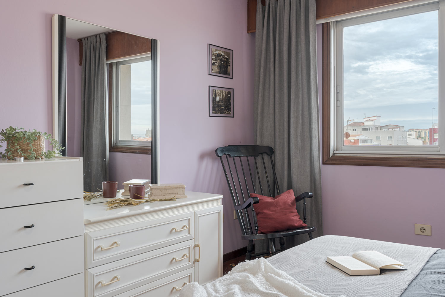 Acogedor rincón de un dormitorio con decoración en tonos lavanda y gris, que incluye un espejo grande, una cómoda blanca, una mecedora negra con cojín rojo y una ventana que ofrece vistas urbanas