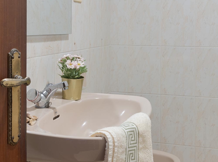 Un baño sencillo con azulejos de mármol beige, un lavabo estándar con una toalla y un pequeño ramo de flores decorativo, creando un ambiente práctico y sin pretensiones