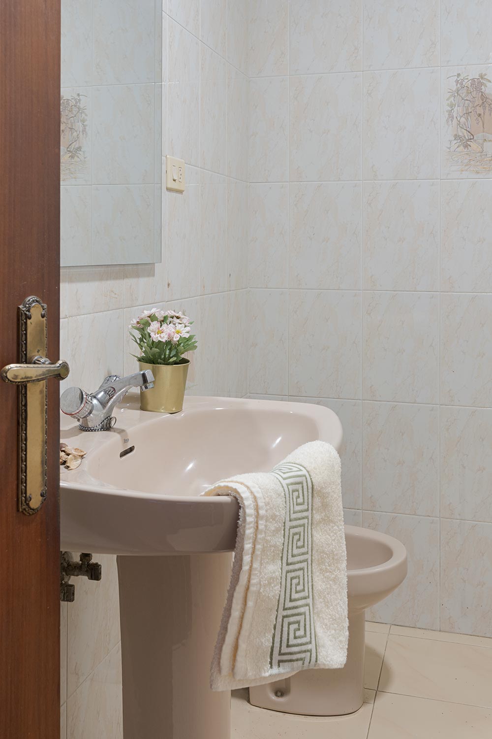 Un baño sencillo con azulejos de mármol beige, un lavabo estándar con una toalla y un pequeño ramo de flores decorativo, creando un ambiente práctico y sin pretensiones