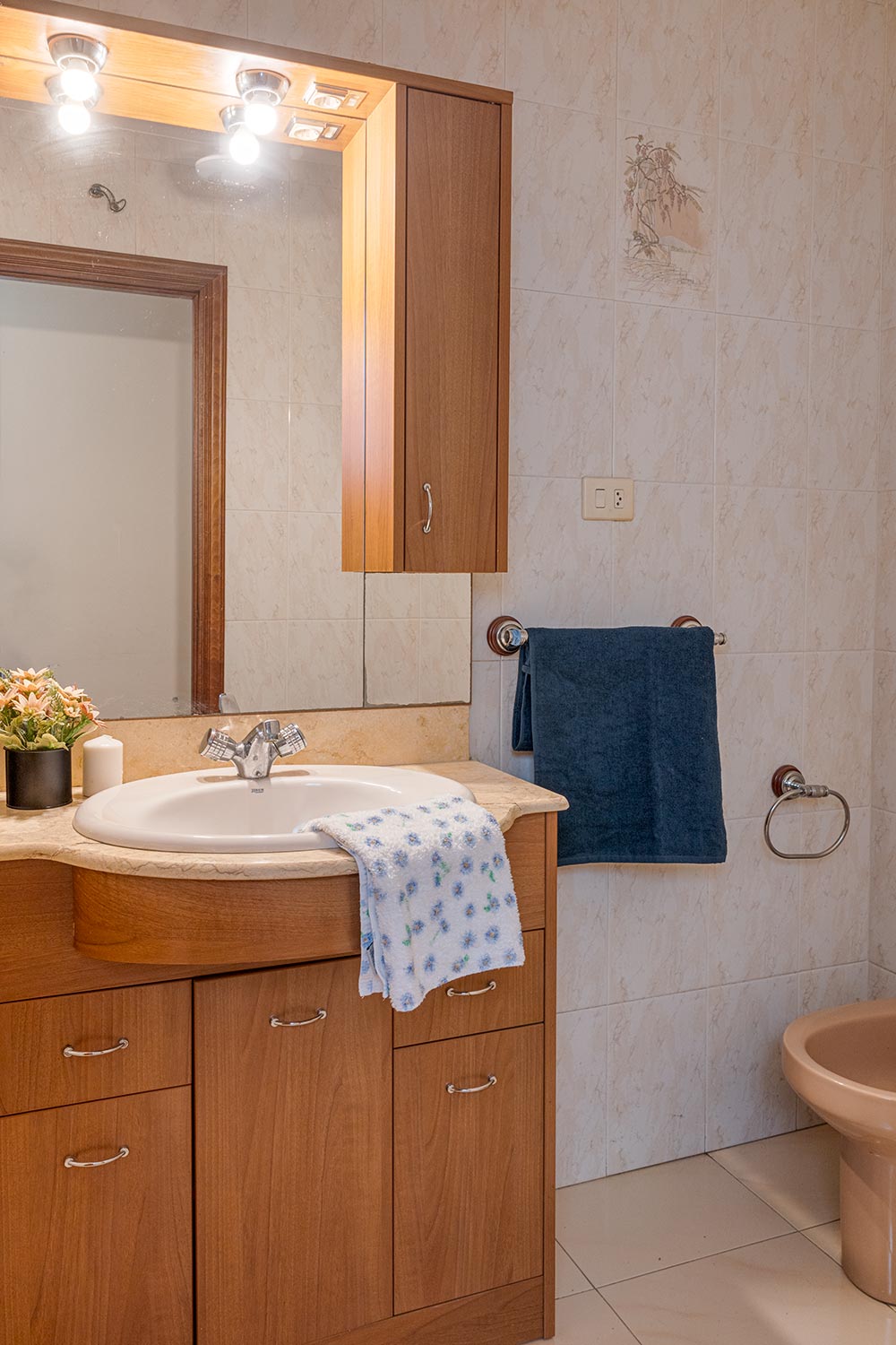 Baño bien iluminado con azulejos claros, mostrando un lavabo con gabinete de madera y un espejo grande con luces en la parte superior. Toallas azules y una planta pequeña adornan la escena, ofreciendo un ambiente práctico y acogedo
