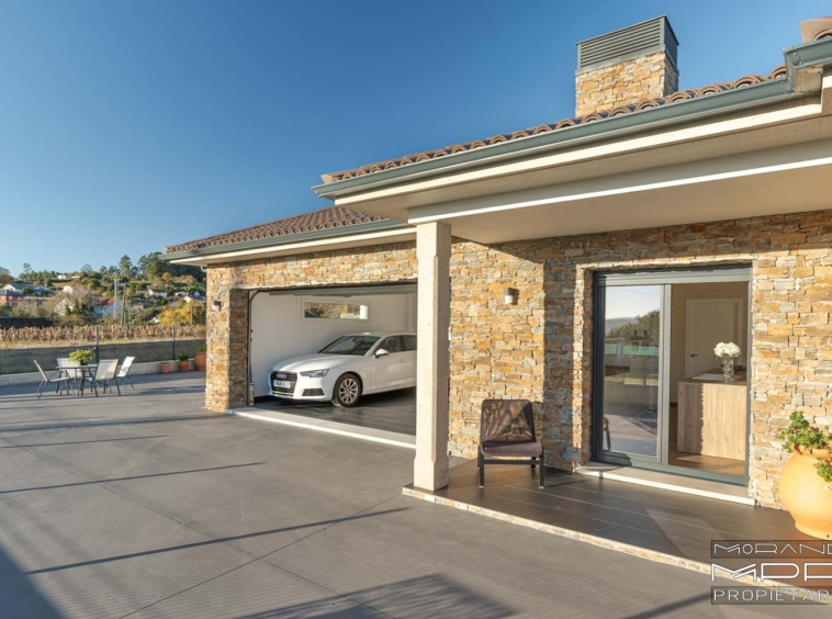 Imagen de una casa moderna de un solo piso con fachada de piedra, garaje abierto con un coche, terraza amplia con mesa y sillas, y cielo despejado.