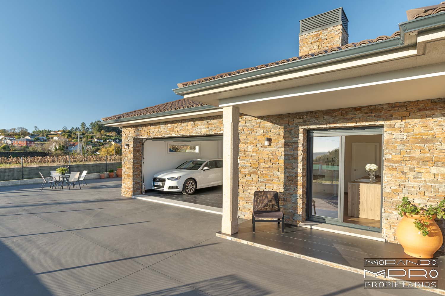 Imagen de una casa moderna de un solo piso con fachada de piedra, garaje abierto con un coche, terraza amplia con mesa y sillas, y cielo despejado.