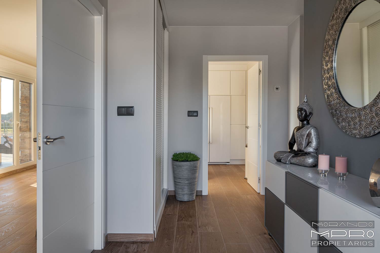 Imagen del interior de una casa, mostrando un pasillo con puertas blancas, suelo de madera y paredes grises. Una figura decorativa de Buda y un gran espejo circular adornan el espacio.