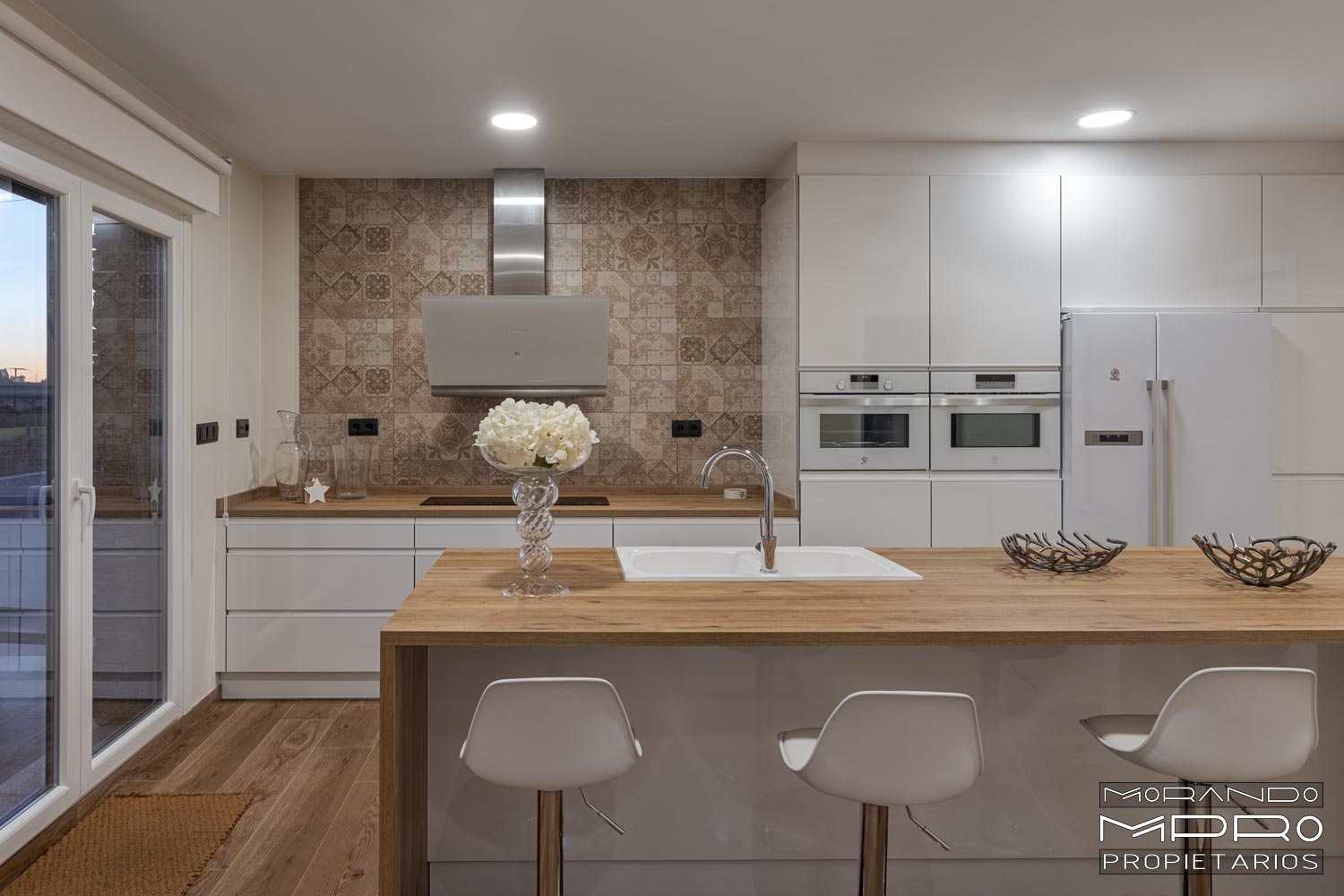 Cocina moderna con muebles en blanco y electrodomésticos a juego con encimera de madera y taburetes blancos.