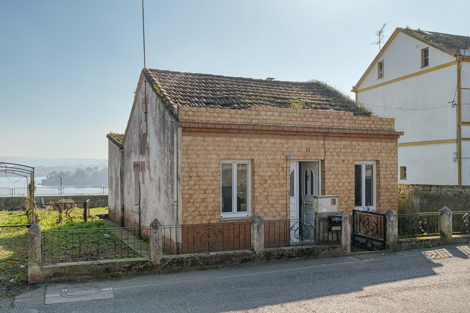 Fachada de una pequeña casa con tejado a dos aguas y vegetación creciendo, ubicada en una tranquila calle de Sada y vistas al mar