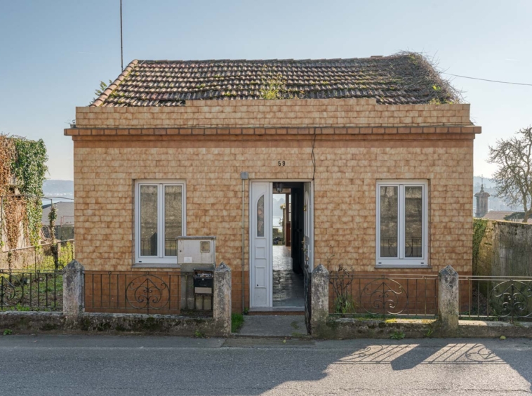 Fachada de una pequeña casa con tejado a dos aguas y vegetación creciendo, ubicada en una tranquila calle de Sada