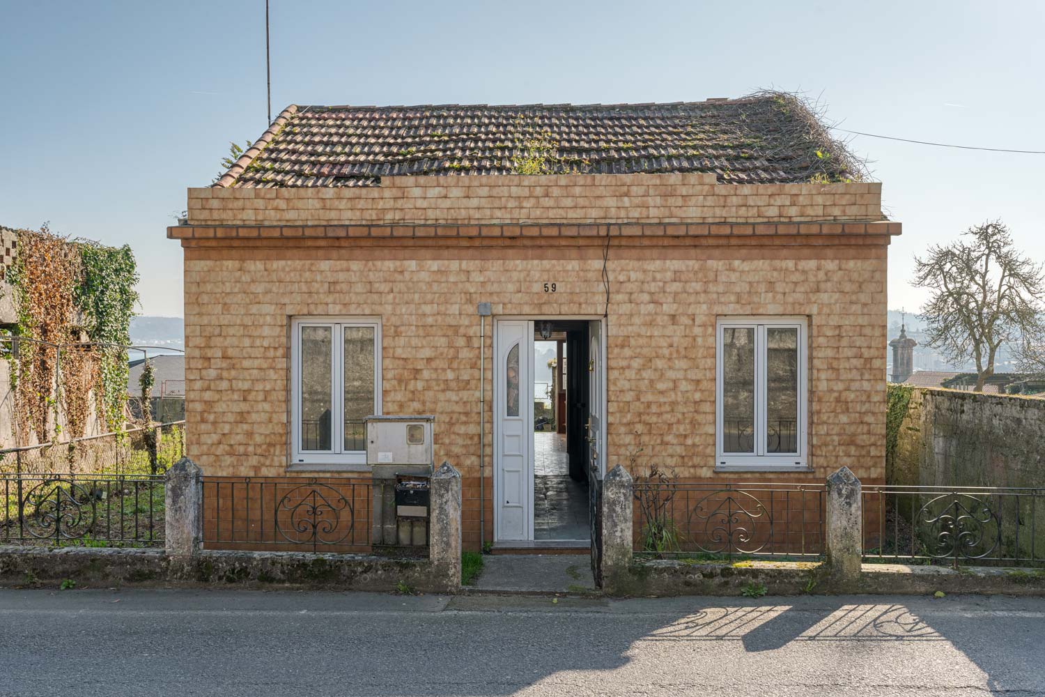Fachada de una pequeña casa con tejado a dos aguas y vegetación creciendo, ubicada en una tranquila calle de Sada