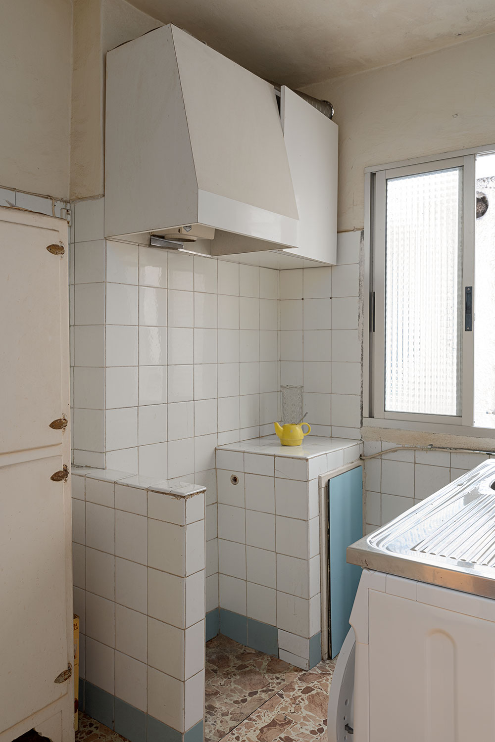 Cocina antigua con azulejos blancos y detalles en azul, equipada con lavadora y ventana pequeña en A Coruña