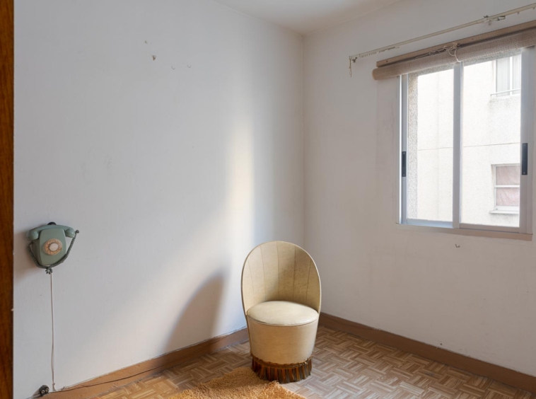 Alt text: "Sencilla habitación con una silla redonda y un teléfono antiguo en la pared, iluminada por la luz natural de la ventana en A Coruña."