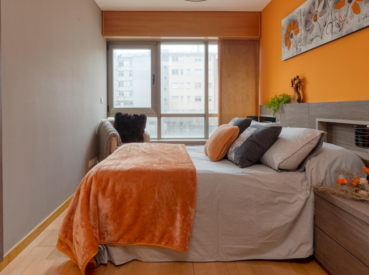 Dormitorio moderno con cama grande, ropa de cama en tonos neutros y cojines naranjas, destacando sobre paredes gris claro y naranja en Sada.