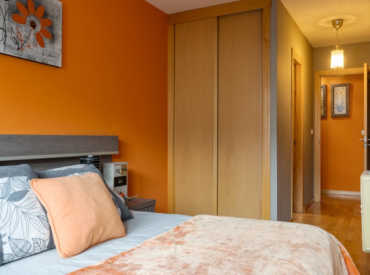 Dormitorio moderno con cama grande, ropa de cama en tonos neutros y cojines naranjas, destacando sobre paredes gris claro y naranja en Sada.