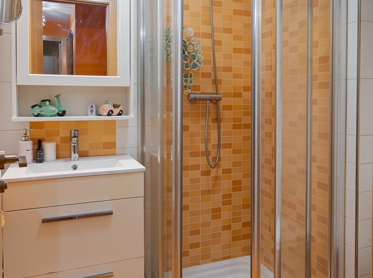 Baño completo con azulejos anaranjados, incluye ducha semicircular con mampara de vidrio, lavabo con mueble blanco y espejo iluminado en Sada