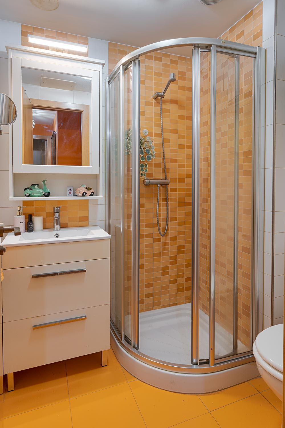 Baño completo con azulejos anaranjados, incluye ducha semicircular con mampara de vidrio, lavabo con mueble blanco y espejo iluminado en Sada