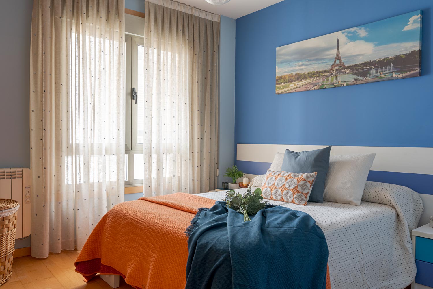Habitación acogedora con paredes azules y franja blanca, cama individual con ropa de cama en tonos naranja y azul, y cortinas claras, en Sada
