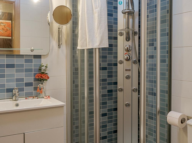 Baño moderno y luminoso con azulejos en tonos azules, ducha de esquina con puertas curvas de vidrio, lavabo blanco con gabinete y espejo circular, en Sada