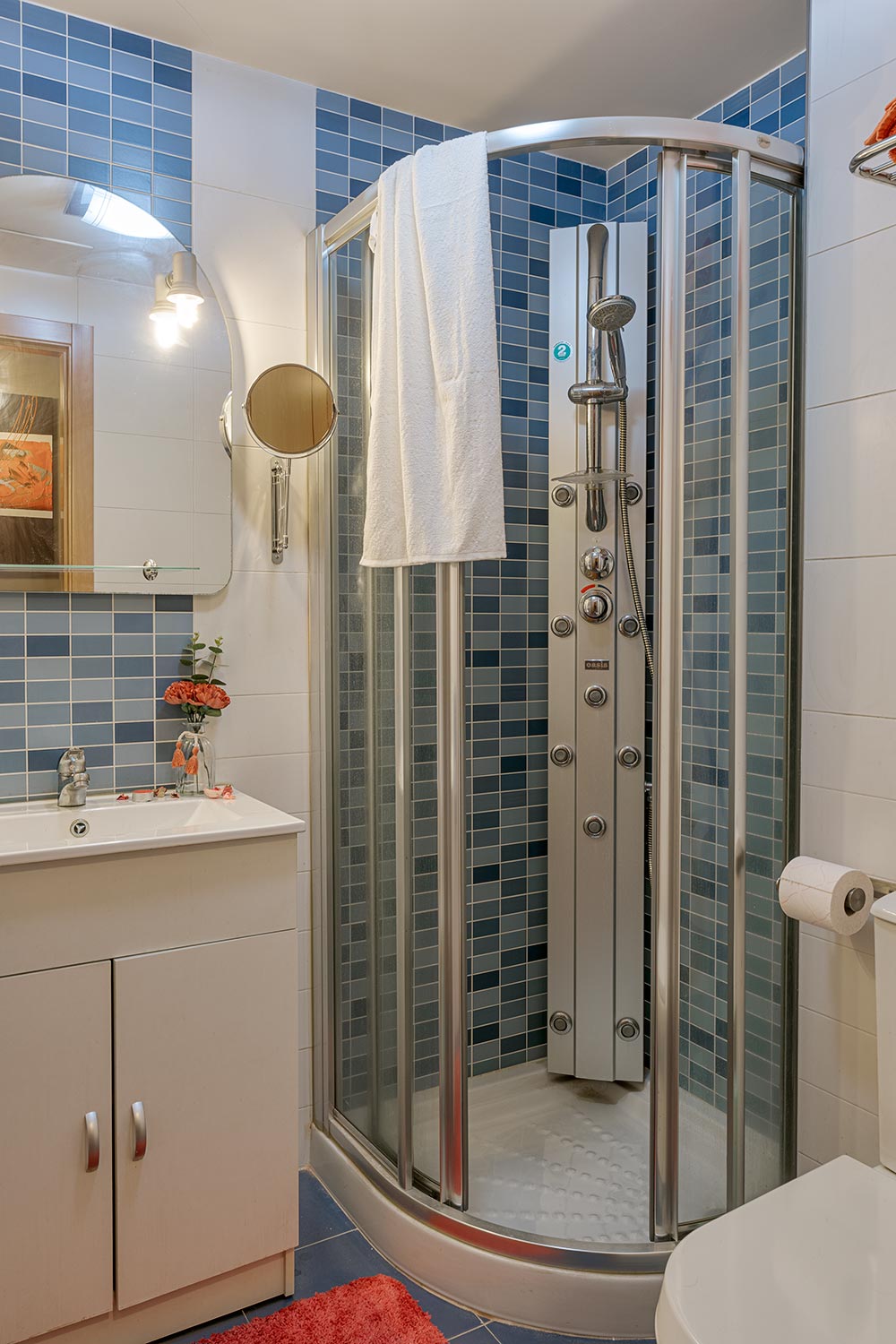 Baño moderno y luminoso con azulejos en tonos azules, ducha de esquina con puertas curvas de vidrio, lavabo blanco con gabinete y espejo circular, en Sada