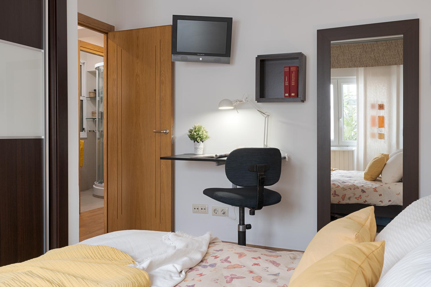 Dormitorio amueblado con una cama con ropa de cama de color neutro, un escritorio con silla, un espejo grande y un televisor montado en la pared.