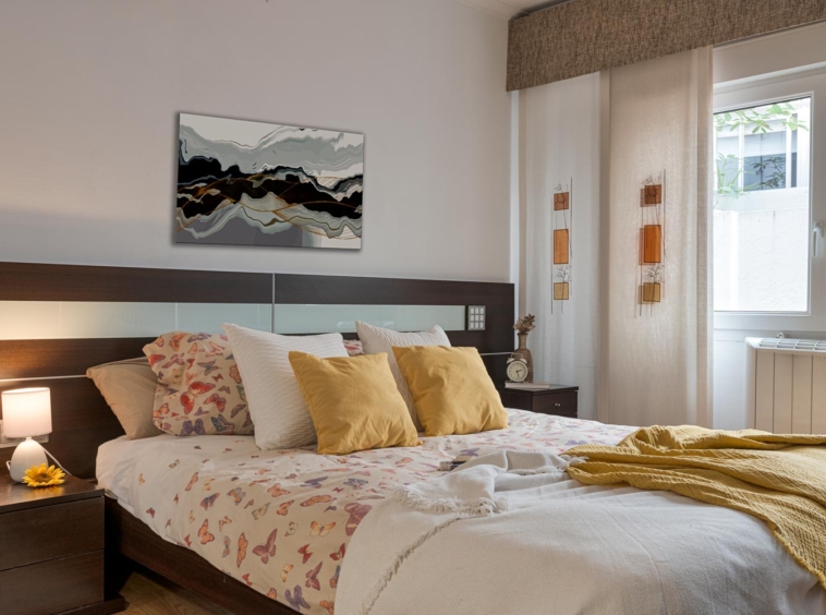 Dormitorio acogedor con una cama de matrimonio con ropa de cama azul, una mesita de noche de madera con una lámpara, un cuadro abstracto en la pared y una ventana con vistas