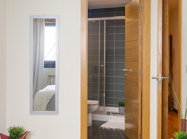 Vista desde una habitación luminosa hacia un baño moderno, destacando la continuidad del suelo de parquet marrón y las puertas de madera clara. El espacio está decorado con espejos, plantas, y una alfombra, creando un ambiente acogedor y hogareño