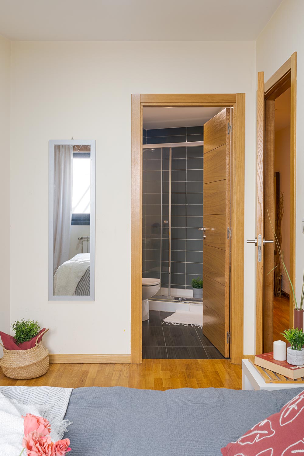 Vista desde una habitación luminosa hacia un baño moderno, destacando la continuidad del suelo de parquet marrón y las puertas de madera clara. El espacio está decorado con espejos, plantas, y una alfombra, creando un ambiente acogedor y hogareño