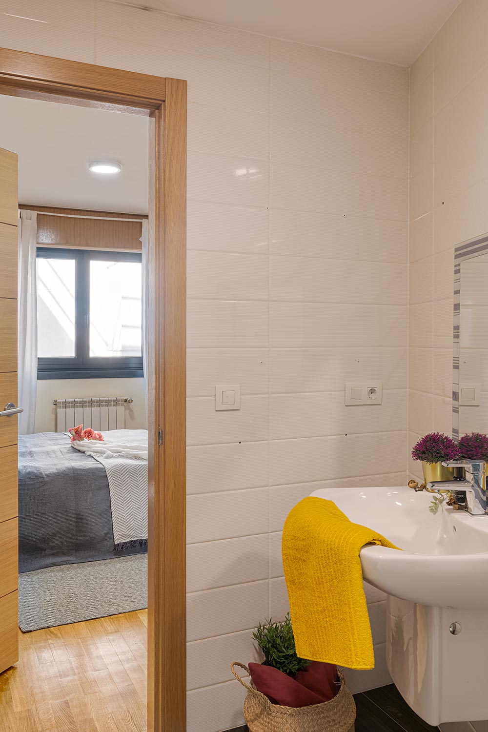 Vista desde el interior de un baño moderno y bien iluminado hacia una habitación acogedora. El baño cuenta con azulejos texturizados en tonos crema, y un lavabo con una toalla amarilla vibrante. En la habitación se ve una cama con ropa de cama gris y una ventana que proporciona luz natural, creando un ambiente hogareño y acogedor.