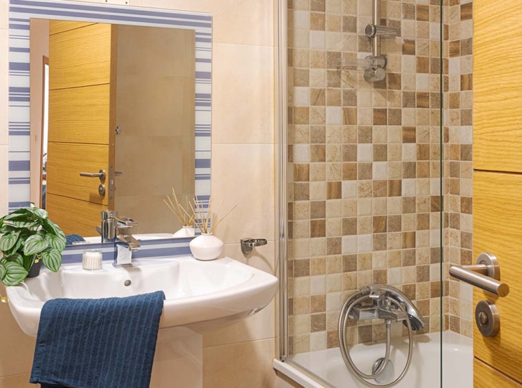 Baño contemporáneo con combinación de azulejos beige y marrones, destacando una bañera con mampara de vidrio y una pared de acento de azulejos a cuadros. Lavabo con espejo amplio y detalles como una toalla azul colgada y una planta verde que añade un toque natural al espacio