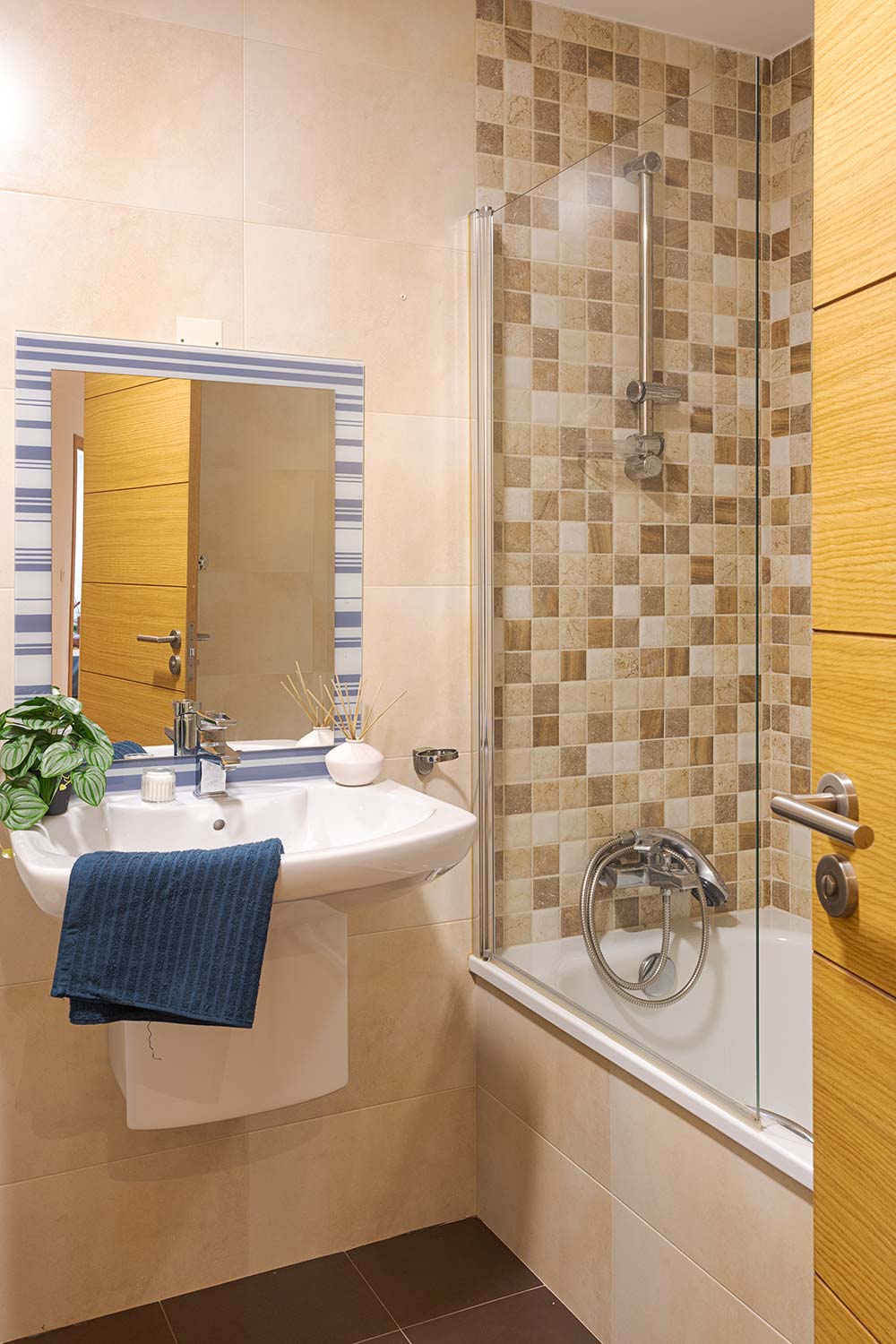 Baño contemporáneo con combinación de azulejos beige y marrones, destacando una bañera con mampara de vidrio y una pared de acento de azulejos a cuadros. Lavabo con espejo amplio y detalles como una toalla azul colgada y una planta verde que añade un toque natural al espacio
